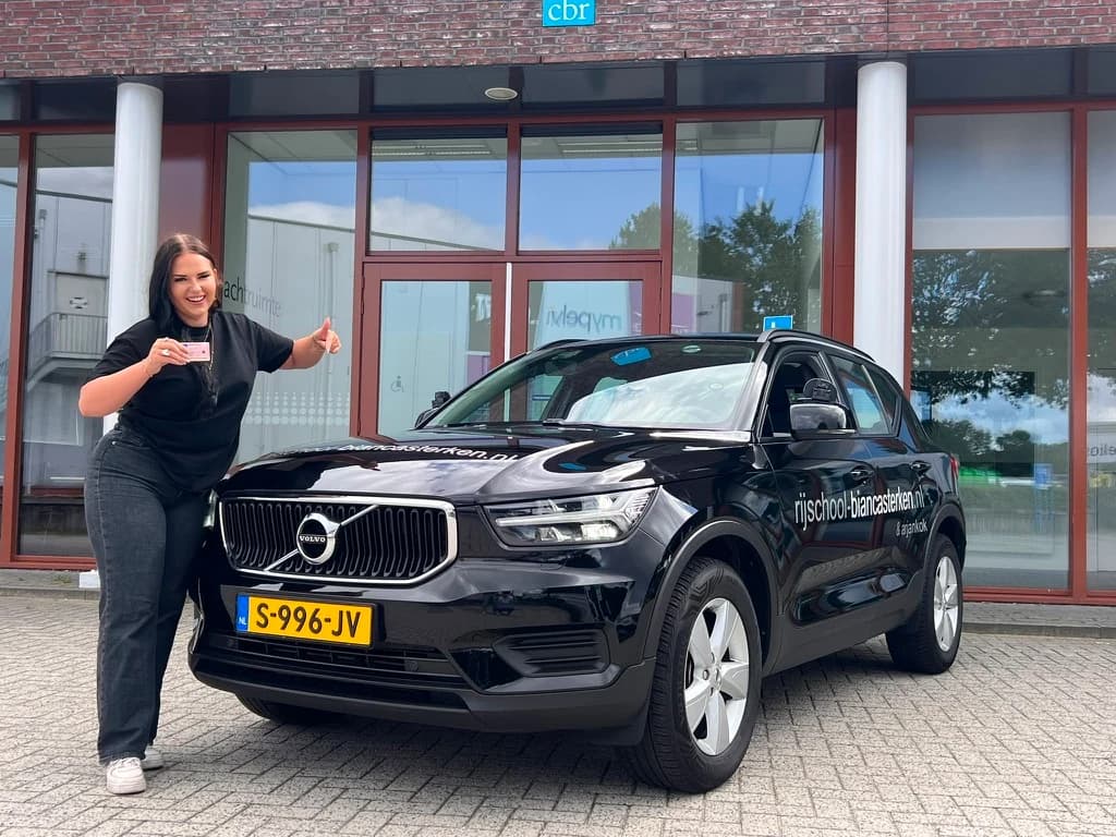 Rijinstructrice Bianca Sterken met een vriendelijke lach naast haar zwarte Volvo lesauto.