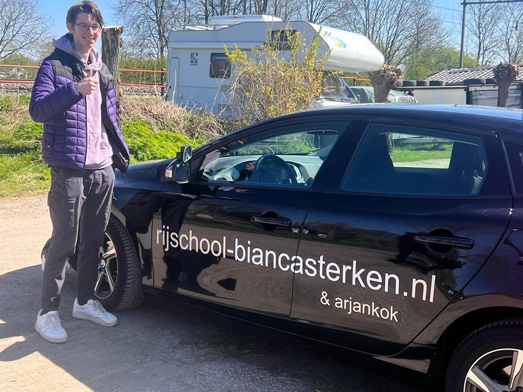 Trotse leerling met duim omhoog bij het portier van de zwarte lesauto.